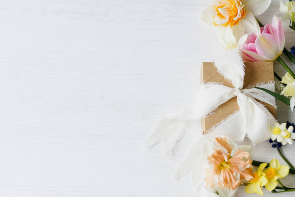 Eleganti fiori primaverili e piatti regalo adagiati su un tavolo bianco rustico, spazio per il testo. Buon giorno delle donne e festa della mamma. Bellissimi narcisi e tulipani bordo bouquet su legno - Foto, immagini