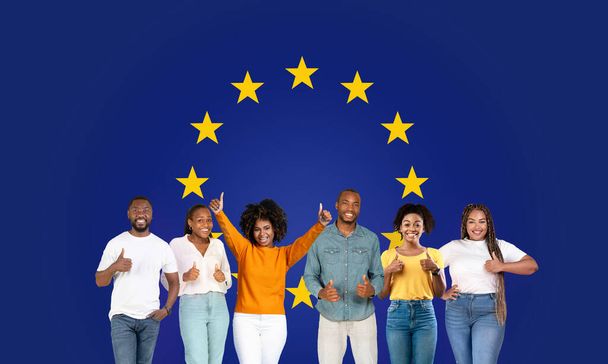 Έξι χαμογελαστοί νεαροί ενήλικες που σηκώνουν τους αντίχειρες, στέκονται με αυτοπεποίθηση με μια έναστρη σημαία της Ευρωπαϊκής Ένωσης στο παρασκήνιο, συνοψίζοντας την ενότητα και τη θετικότητα, συστήνουν, στούντιο, πανόραμα - Φωτογραφία, εικόνα