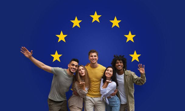 Cinq amis exubérants milléniaux embrassant et agitant avec joie, debout ensemble sur un fond bleu profond orné des étoiles dorées, studio, panorama de l'Union européenne - Photo, image