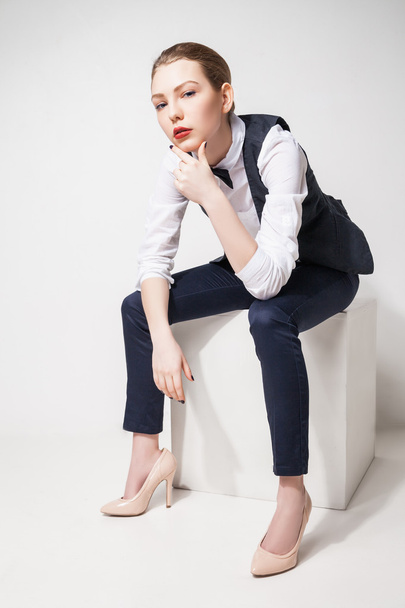 fashion model in black trousers, top, bow tie and vest sitting on cube over white - Foto, Imagem