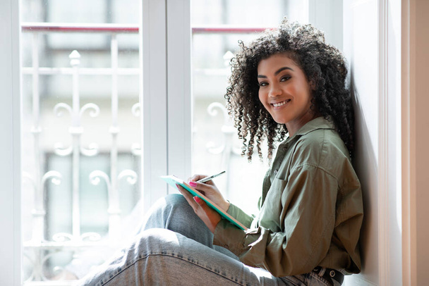 Happy Black College student dame zit bij het raam thuis, met notitieblok en notities, geniet van het weekend van leren en planning binnen, glimlachen om de camera. Concept van creatief schrijven - Foto, afbeelding