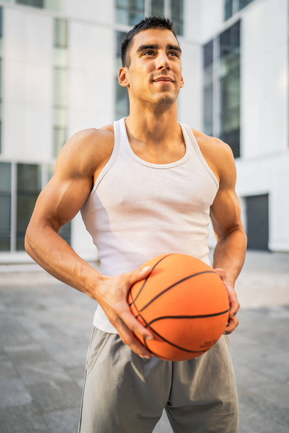 Un joven hombre caucásico atleta masculino de pie al aire libre mantenga la pelota de baloncesto usar blanco tanque superior una camisa fuerte muscular persona real copiar espacio sonrisa feliz confianza estilo de vida saludable concepto - Foto, imagen