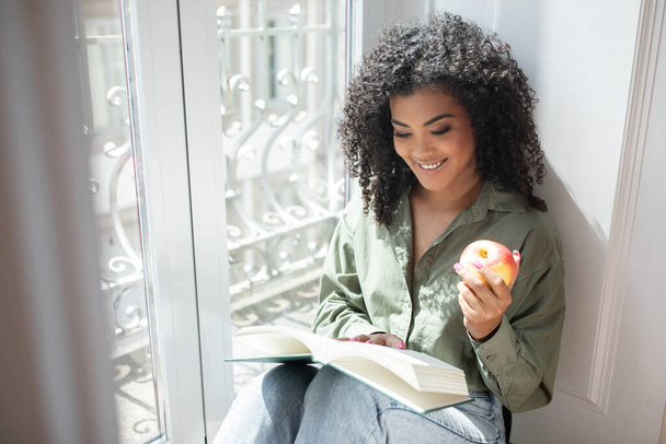 Freude am Lesen. Die glückliche Brasilianerin schwelgt in einem Bestseller, liest am Fenster mit Apfelfrüchten und genießt ihr entspanntes Wochenende im gemütlichen Heim. Kopierraum - Foto, Bild