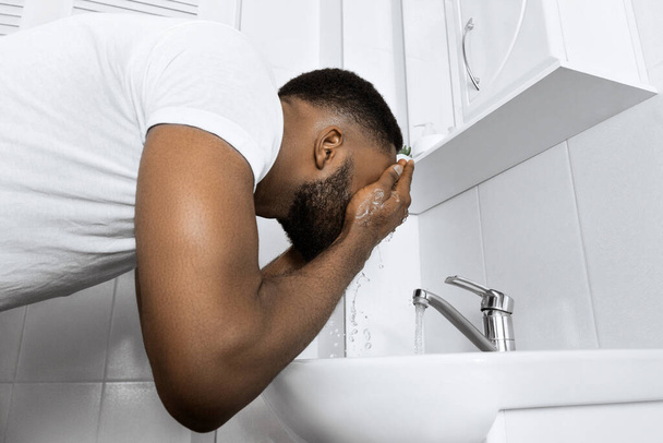 Молодий афро-чоловік миє обличчя в раковині у ванній кімнаті, копіювати простір
 - Фото, зображення