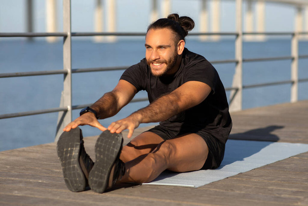 Boldog atlétikai srác a sportruházat bemutatja ülő előre kanyar a szőnyegen, edz a tengerparti szabadban, bemutatja a rugalmasság és elősegíti a testmozgás a jólét és a fit életmód - Fotó, kép