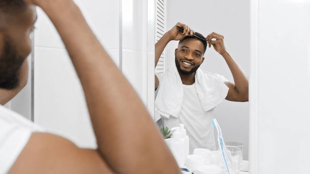 ミレニアル世代のアフロ男は髪をコーミングし、バスルームで鏡を見て。現代人のコンセプトの朝 - 写真・画像