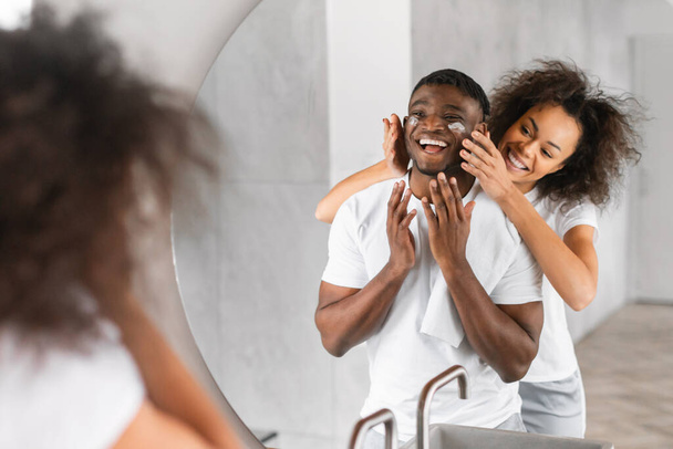 African American αγαπώντας ζευγάρι επιδίδεται σε κοινή ρουτίνα φροντίδας του δέρματος στο μπάνιο, καθώς η σύζυγος εφαρμόζει ενυδατική κρέμα στο δέρμα του συζύγου. Αγάπη και σύνδεση μεταξύ ζεύγους χιλιετιών, οικογένεια περιποίηση - Φωτογραφία, εικόνα