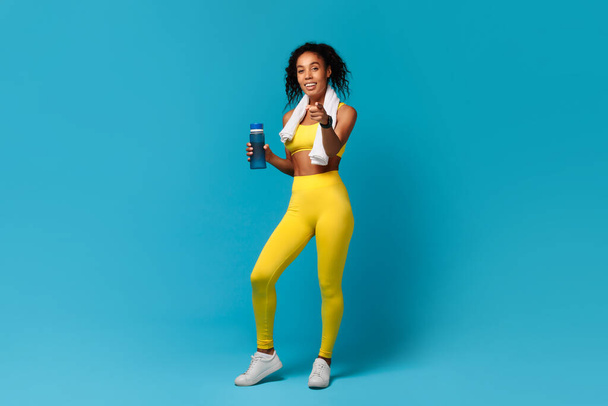 Aktiver Lebensstil. Lächelnde athletische schwarze Frau in gelber Sportbekleidung hält Wasserflasche in der Hand, zeigt mit dem Finger in die Kamera, motiviert zum gesunden Abnehmen und Workout, steht auf blauem Studiohintergrund - Foto, Bild