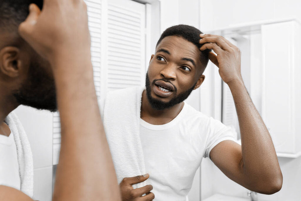 Millennial Afro człowiek dotykając jego włosy, frown i patrząc w lustro w łazience. Koncepcja problemu włosów i rzeźb - Zdjęcie, obraz