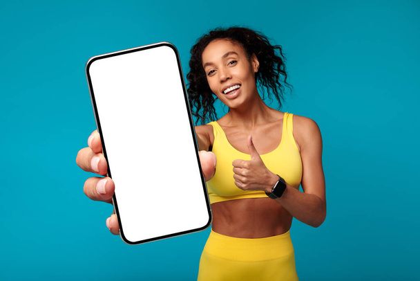 黄色いスポーツウェアの幸せなアフリカ系アメリカ人女性は,彼女のスマートフォンが空の画面を示し,青いスタジオの背景に動機と承認を表現しながら,親指を与えます. フィットネスアプリのモックアップ - 写真・画像