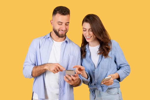 Χαρούμενος άνδρας και γυναίκα σε περιστασιακή ανταλλαγή ρούχων συναρπαστικό περιεχόμενο στα τηλέφωνά τους, απολαμβάνοντας τη στιγμή της ψηφιακής αλληλεπίδρασης σε ζεστό κίτρινο φόντο - Φωτογραφία, εικόνα