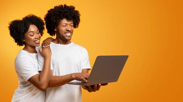 ポジティブなミレニアル世代のアフリカ系アメリカ人男性と女性カップルは,オレンジ色のスタジオの背景,ウェブサーフィン,映画を見る,コピースペースを持つパノラマを使用してラップトップコンピュータを使用して白いTシャツを着ています - 写真・画像