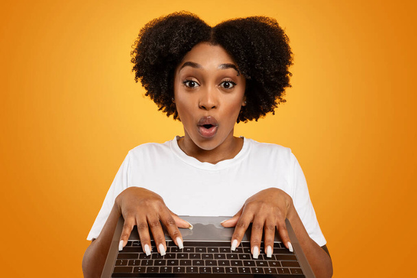 Amazed hübsche junge afrikanisch-amerikanische Dame tippt auf Laptop-Computer-Tastatur, Grimassen, Websurfen, Überprüfung großer Online-Angebot, orangefarbener Hintergrund, Nahaufnahme - Foto, Bild