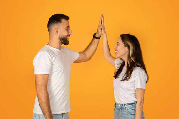 Glückliches junges Paar, das eine High-Five gibt und den Erfolg mit freudigem Gesichtsausdruck feiert. Beide tragen weiße T-Shirts und blaue Jeans vor orangefarbenem Hintergrund und symbolisieren Teamwork und Leistung. - Foto, Bild