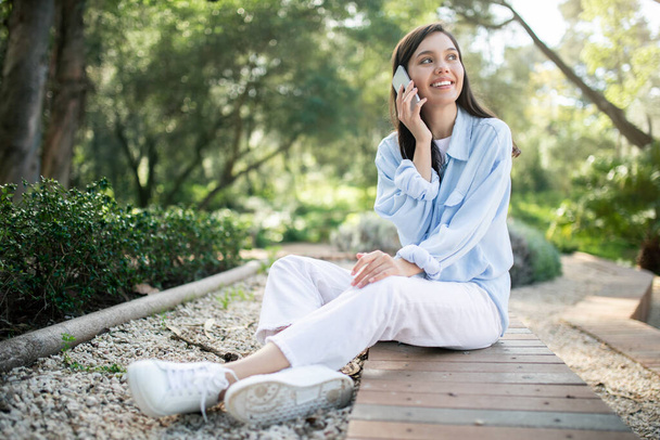 Uma jovem caucasiana alegre em traje casual desfruta de uma conversa agradável em seu smartphone, sentada em um caminho de madeira em um parque exuberante, comprimento total, chamada na cidade, ao ar livre - Foto, Imagem