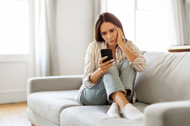 Mujer joven molesta sentada en el sofá y mirando su teléfono inteligente con expresión reflexiva o preocupada, mujer milenaria reaccionando a noticias o mensajes inesperados mientras se relaja en el sofá en casa - Foto, imagen