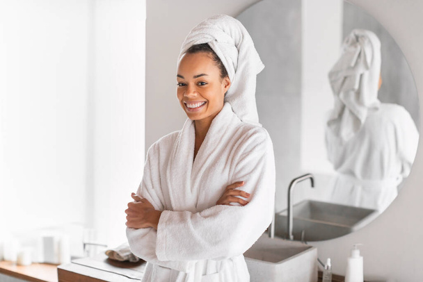 Napi szépség rutin. Afrikai amerikai Y-generációs hölgy áll összecsukott karokkal fürdőköpenyben, élvezi a reggeli önellátó rituálékat, mosolyog a kamerába a modern fürdőszobában. Kényeztetés és wellness - Fotó, kép