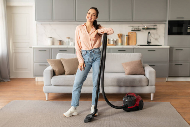 Confident, smiling woman in pastel blouse and jeans poses with vacuum cleaner in chic, well-kept living space, exuding casual elegance, full length - 写真・画像