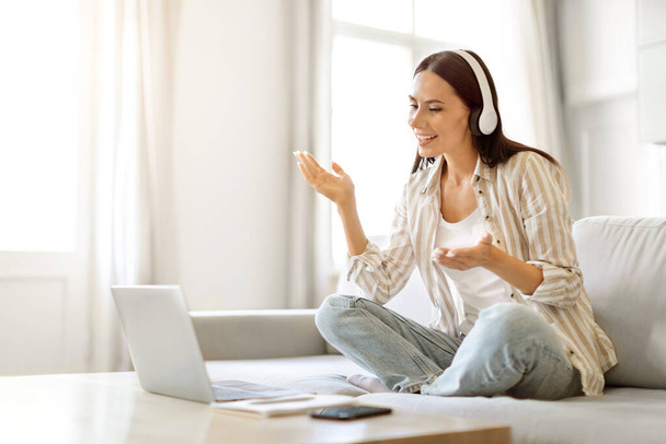 Відеодзвінок. Щаслива молода жінка в навушниках Телеконференція на ноутбуці вдома, посміхаючись Кавказька жінка говорить і жестикулює на комп'ютерній веб-камері, сидячи на дивані, копіювати простір - Фото, зображення