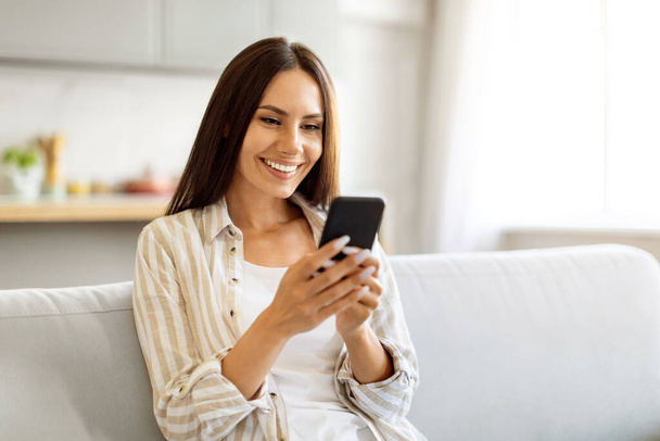 Uśmiechnięta piękna kobieta wpisując wiadomość na smartfonie siedząc na kanapie w domu, szczęśliwa młoda Europejka korzystająca z telefonu komórkowego do komunikacji lub zakupów online, skopiuj przestrzeń - Zdjęcie, obraz