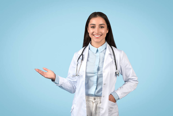 Gülümseyen kadın doktor tek eliyle görünmez bir şey sunuyor, steteskop ve beyaz laboratuvar önlüğü giyiyor, sakin mavi arka plana karşı güvenle duruyor. - Fotoğraf, Görsel