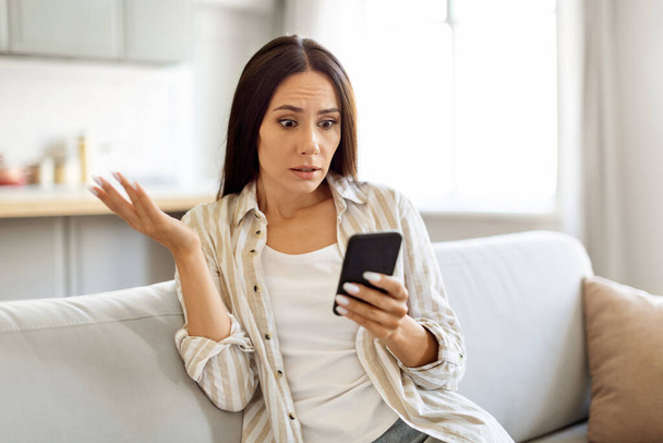 Geschokt jonge vrouw lezen van slechte boodschap op de smartphone tijdens het zitten op de bank thuis, Verward Millennial vrouw met mobiele telefoon in handen Emotioneel reageren op onaangenaam nieuws, Kopiëren ruimte - Foto, afbeelding