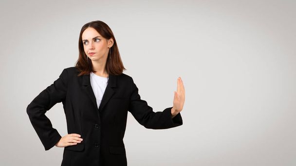 Mulher de negócios millennial caucasiana desconfiada em uma jaqueta de terno preto mostrando um sinal de mão parada, com uma expressão cética, indicando cautela ou recusa, contra um fundo simples - Foto, Imagem