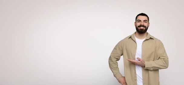 Cheerful man with a beard presenting with his hand to the side, displaying an empty space for text or product, wearing a beige shirt and a white t-shirt on a wide background - Photo, Image
