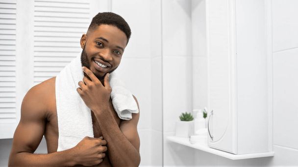 Smiling afro man with naked muscular torso touching his beard at bathroom. Mans daily care concept, copy space - Photo, Image