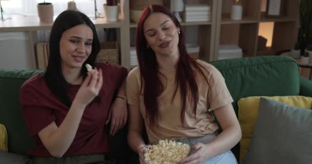 dos mujeres caucásicas adolescentes amigos o hermanas ver película serie de televisión en casa sentarse en el sofá cama con palomitas de maíz - Metraje, vídeo