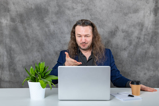 Un hombre profesional enfocado con el pelo largo está sentado en un escritorio en una oficina contemporánea, trabajando diligentemente en un ordenador portátil, inmerso en sus tareas y rodeado por un entorno de oficina moderno. - Foto, Imagen