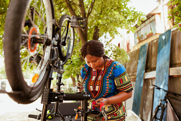 Energetyczna afrykańska amerkańska rowerzystka uważnie bada specjalistyczny sprzęt do serwisowania rowerów na zewnątrz. Sport miłośnik czarny kobieta szuka narzędzia pracy w domu podwórko dedykowane do lata - Zdjęcie, obraz
