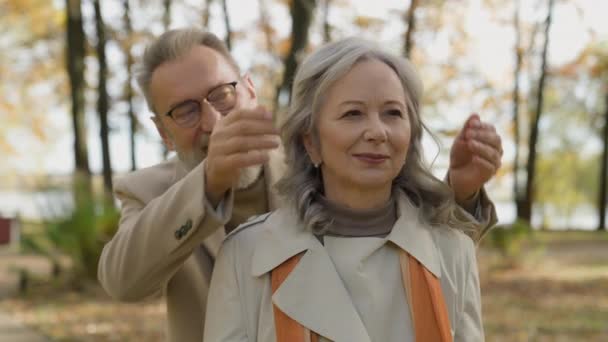 Senior gray-haired man closing woman eyes with hands make surprise in autumn park outdoors Caucasian couple mature beautiful female wife retired male husband happy old aged family romantic feelings - Footage, Video