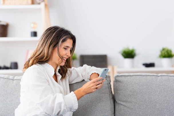 Sivunäkymä kypsä nainen käyttää mobiili istuu sohvalla kotona - Valokuva, kuva