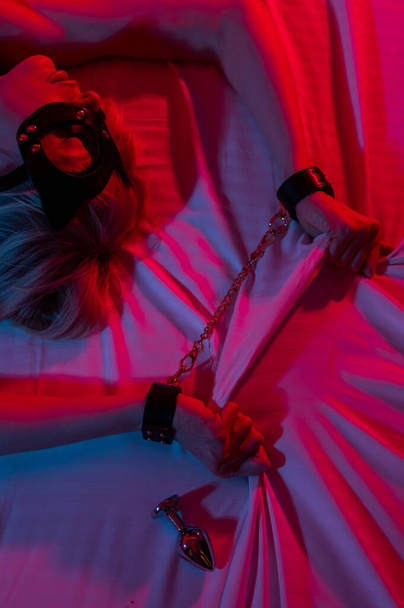 Sexy blonde woman in a leather mask and handcuffs and a collar lies in the bedroom in a red-blue neon light. Role-playing games - Foto, afbeelding