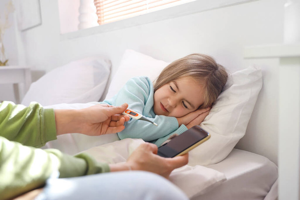 Sick little girl with her mother holding thermometer and mobile phone in bedroom - Photo, Image