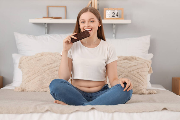 Happy young woman in tight jeans eating chocolate at home. Weight gain concept - Photo, Image