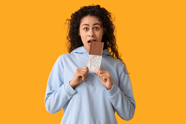 Mooie jonge Afro-Amerikaanse vrouw eten zoete chocoladereep op gele achtergrond - Foto, afbeelding