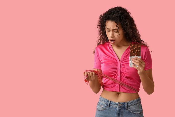Jeune femme afro-américaine choquée avec barre de chocolat sucrée et ruban à mesurer sur fond rose - Photo, image