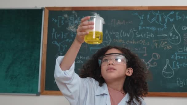 Atraktivní americký student drží kádinku a kontroluje biochemické řešení. Detailní záběr zvědavé dívky míchání kádinka a při pohledu na změnu barvy, zatímco stojí v laboratoři s tabulí. Pedagogika. - Záběry, video