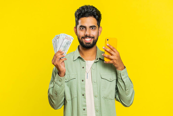 Indian man looking smartphone display sincerely rejoicing win, receiving money dollar cash banknotes celebrating success victory play lottery game. Lucky rich Arabian guy isolated on yellow background - Photo, Image