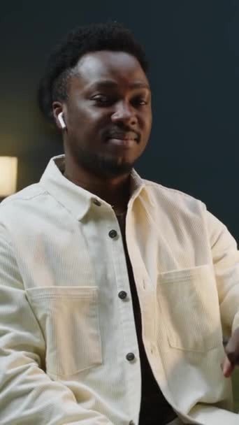 Függőleges közepes lassú portré fiatal magabiztos afro-amerikai üzletember vezeték nélküli fülhallgató pózol kamera, miközben ül az íróasztal mellett a kortárs irodában - Felvétel, videó