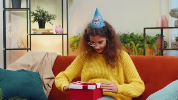 ハッピー14-15歳の子供の少女は,お祝いの誕生日帽子の帽子のホールドギフトボックスを着て,リボンを祝福します. ティーンエイジャー 女性 子供 お祝い パーティー イベント オープニング 配信 ソファーの自宅の部屋でプレゼント - 映像、動画