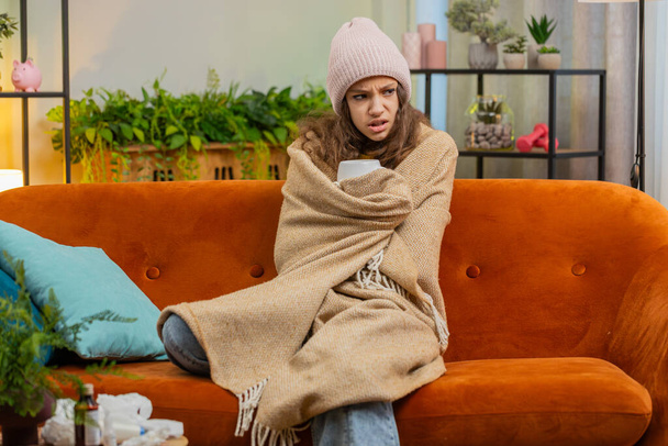 病気の子供の少女は,重い負債を加えることなく,暖房されていない家のアパートで熱いお茶を飲むソファーで冷たい風邪から単独で座って包まれた帽子を着ます. 不健康な子供は不快感を感じる 温暖化しようとする - 写真・画像
