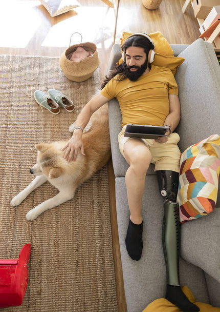 Biyonik bacaklı adam, köpeği ile kanepeye uzanıp dijital tablette film izliyor. - Fotoğraf, Görsel