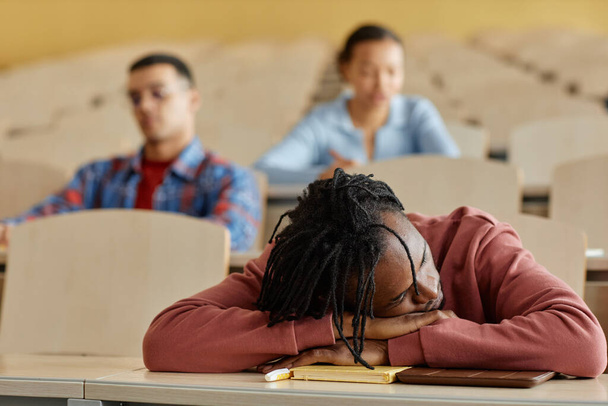 Afrikai amerikai iskolás fiú fekszik az asztalon, és alszik közben unalmas előadás az osztálytársai a háttérben - Fotó, kép