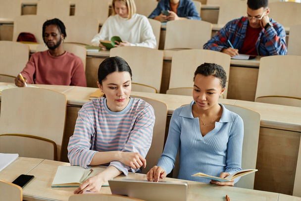 Дві школярки використовують ноутбук разом, сидячи за столом під час лекцій з іншими студентами на задньому плані - Фото, зображення
