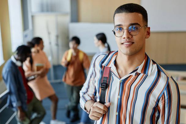 Портрет студента в очках с рюкзаком, улыбающегося перед камерой, стоящей в лекционном зале колледжа - Фото, изображение