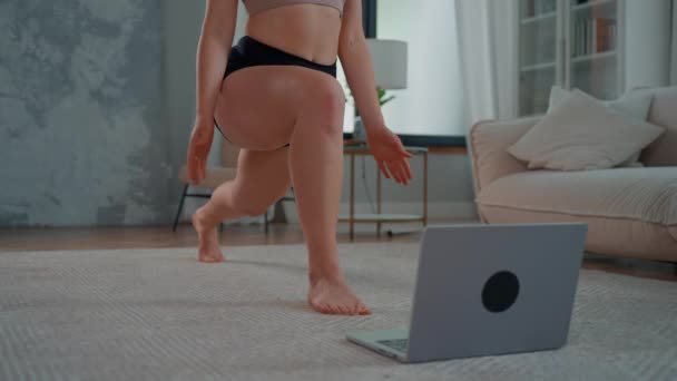 Μια γυναίκα φορώντας σορτς και τοπ κάνει γιόγκα κατάρτισης με τον προπονητή online βλέποντας στην οθόνη laptop επανάληψη άσκηση γυμναστικής εσωτερική  - Πλάνα, βίντεο