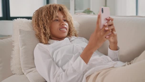 女性は携帯電話を持っている間にソファーに横たわっているのを見かけます. 彼女はリラックスして,電話の画面に焦点を当てているようです.. - 映像、動画